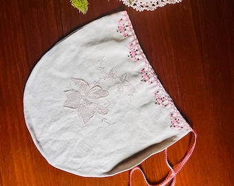 Puede incluir: Un gorro blanco con delicados bordados florales y un borde de cinta rosa. El gorro incluye un borde decorativo con detalles tejidos en rosa y dos finas cintas rosas en la parte inferior, cada una terminada con una pequeña cuenta azul.