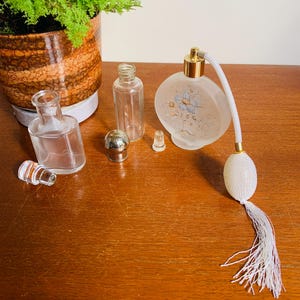 May include: A set of vintage glass perfume bottles with a frosted floral design. The set includes a round bottle with a spray nozzle and a tassel, a small bottle with a stopper, and a clear bottle with a stopper. The bottles are on a wooden surface.