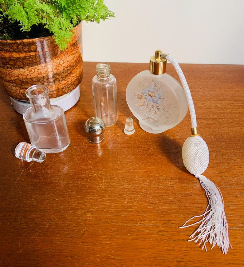 May include: A set of vintage glass perfume bottles with a frosted floral design. The set includes a round bottle with a spray nozzle and a tassel, a small bottle with a stopper, and a clear bottle with a stopper. The bottles are on a wooden surface.