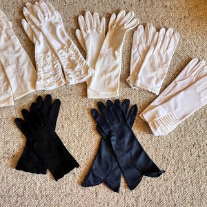 May include: A collection of seven pairs of gloves in various styles, including white pairs with ruffled cuffs, lace trim, and embroidered details, alongside two black pairs featuring scalloped edges and intricate floral embroidery on the wrists.