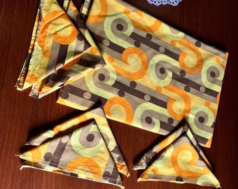 May include: A rectangular placemat and three folded napkins featuring a repeating pattern of orange, yellow, and brown circular swirls and horizontal stripes. The fabric items are arranged on a dark wood surface with a green plant visible.