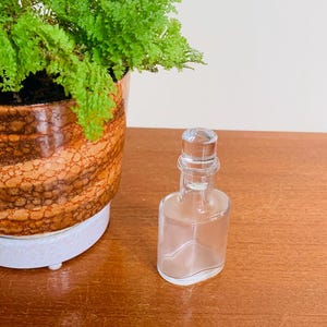 May include: A clear glass bottle with a glass stopper. The bottle is empty and has a rounded rectangular shape. The bottle is sitting on a wooden surface.