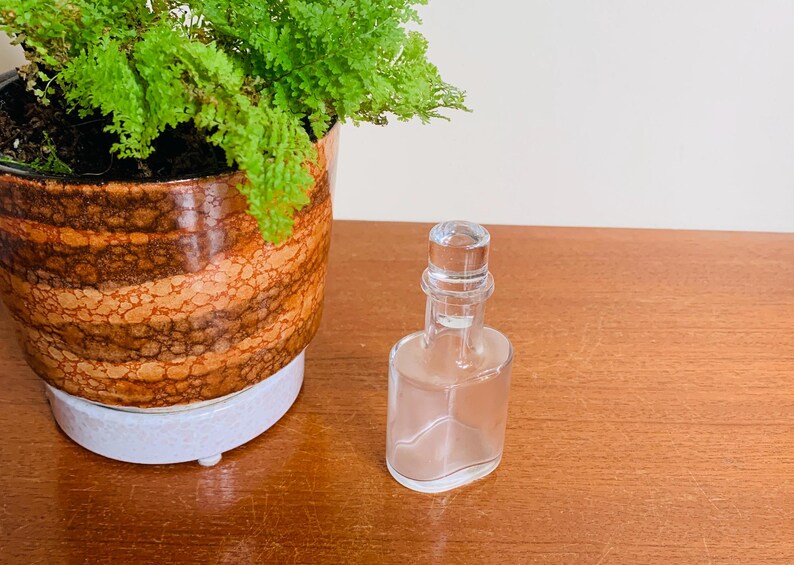 May include: A clear glass bottle with a glass stopper. The bottle is empty and has a rounded rectangular shape. The bottle is sitting on a wooden surface.