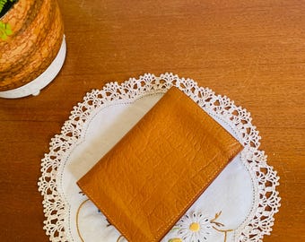 Puede incluir: Una cartera rectangular de cuero color canela con una superficie texturizada y en relieve. La cartera está plegada y presenta costuras visibles a lo largo de los bordes, mostrando un diseño estructurado con un tono marrón cálido y uniforme.