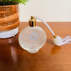 May include: A clear glass perfume bottle with a gold top and a white floral design. The bottle has a white tassel attached to the top.