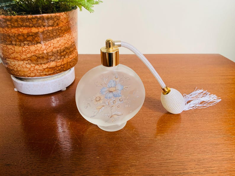 May include: A clear glass perfume bottle with a gold top and a white floral design. The bottle has a white tassel attached to the top.