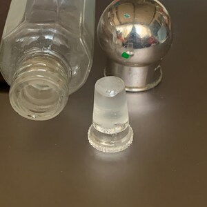 May include: A clear glass bottle with a silver metal stopper and a clear glass dropper. The bottle has a hexagonal shape and a textured surface.