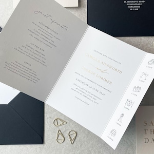 May include: Elegant wedding invitation suite featuring a tri-fold invitation with gold lettering and icons, a dark navy envelope, and gold paper clips. The invitation includes the names Camilla Ainsworth and George Lorimer, and the date.