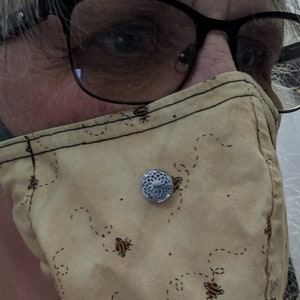 Puede incluir: Una persona lleva una mascarilla beige con un patrón de abejas y un botón plateado. La mascarilla tiene un borde cosido negro y una correa negra. La persona también lleva gafas con montura negra.