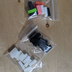 May include: Three small plastic bags filled with different colored foam pieces. The bags are on a light brown wooden surface. The foam pieces are in various shapes and sizes.