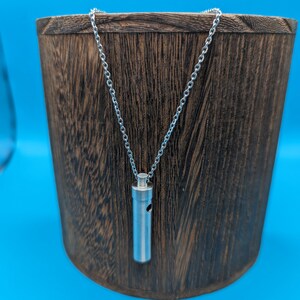 Puede incluir: Un collar de cadena de plata con un colgante cilíndrico. El colgante tiene un pequeño agujero en el lateral. El collar se muestra sobre una superficie de madera.