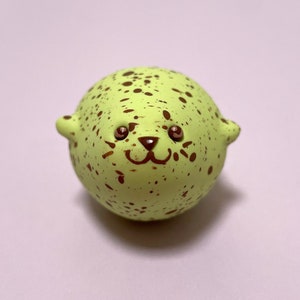 以下が含まれることがあります： かわいい顔をした、小さく丸い緑と茶色のアザラシの置物。アザラシは茶色の目と茶色の鼻をしていて、茶色の斑点で覆われています。