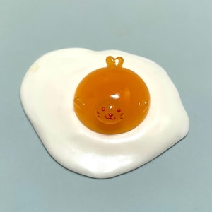 May include: A small, smooth, resin-like figurine shaped like a fried egg. The white portion forms an irregular, flat base, while the centre features a raised, translucent orange yolk with a moulded seal face, including tiny red eyes and whiskers.