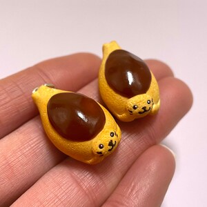 Könnte beinhalten: Zwei braune und gelbe Tonanhänger in Form eines Brötchens mit brauner Glasur oben. Jeder Anhänger hat ein süßes Comic-Gesicht mit schwarzen Augen und einem Lächeln.