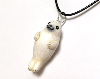 May include: A handmade white seal pendant necklace with a black cord. The seal charm has black eyes and a smiling mouth. The pendant is attached to a silver-colored ring.