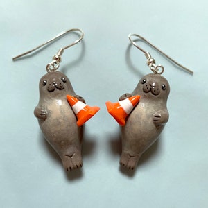Op de afbeelding: Een paar zilveren oorbellen met een grijze zeehond die een oranje verkeerskegel vasthoudt. De oorbellen zijn gemaakt van polymer clay en zijn een leuke en grillige accessoire.