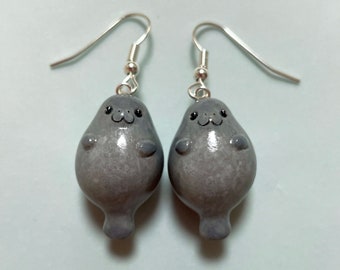 May include: A pair of silver dangle earrings featuring gray, hand-painted, polymer clay sea lion charms. The sea lions have black eyes and a cute, cartoon-like expression.