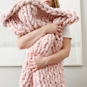 May include: A soft, pink, chunky knit blanket is being held by a person. The blanket is made of thick yarn and has a fluffy texture.