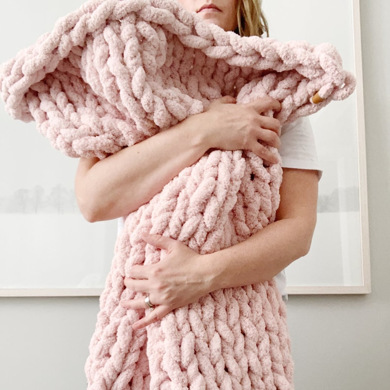 May include: A soft, pink, chunky knit blanket is being held by a person. The blanket is made of thick yarn and has a fluffy texture.