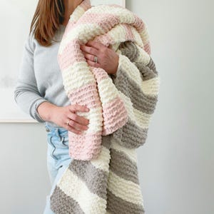 Peut inclure: Une couverture en tricot épais avec un motif rayé rose, blanc et gris. La couverture est tenue, mettant en valeur sa texture épaisse et sa palette de couleurs. Cette couverture douillette et faite à la main est un article de décoration intérieure.