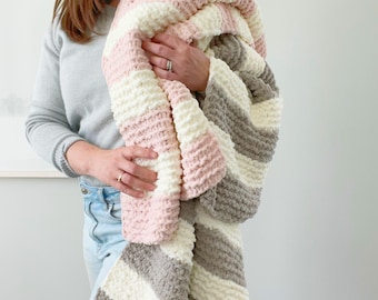 May include: A chunky knit blanket with a striped design in pink, white, and grey. The blanket is held up, displaying its thick texture and colour scheme. This cosy, handmade blanket is a home decor item.