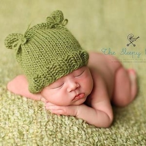 May include: A sleeping baby wearing a green knitted hat with two small pom-poms on top. The hat has a ribbed texture and is pulled down over the baby's forehead.