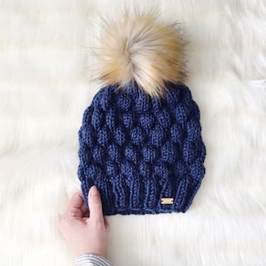 May include: A navy blue knitted beanie with a fluffy brown pom pom. The beanie has a textured, bubble-like design.