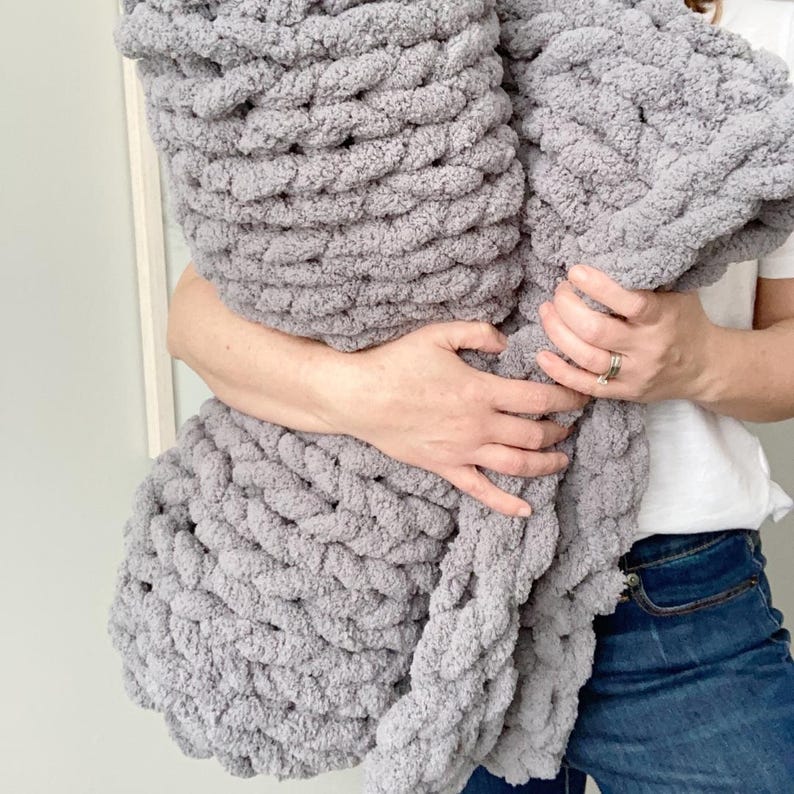 May include: A soft, grey chunky knit blanket is held in the arms of a person. The blanket is made with thick yarn and has a fluffy texture.