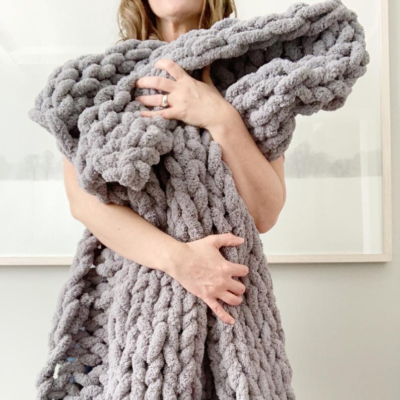 May include: A grey chunky knit throw blanket with a cable knit pattern. The blanket is being held by a person.