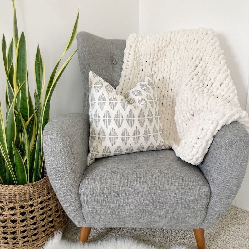 May include: A grey armchair with a white chunky knit throw blanket and a grey and white geometric patterned cushion. A green snake plant is in a woven basket to the left of the chair.