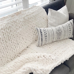 May include: A white chunky knit throw blanket with a striped and patterned pillow on top of it.