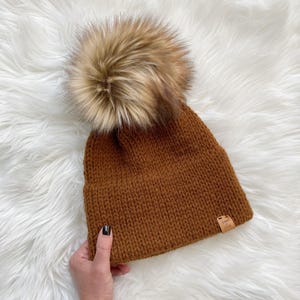 May include: A brown knitted beanie with a large faux fur pom pom on top. The beanie has a small brown leather tag with the text "Cozy & Chic" on it.
