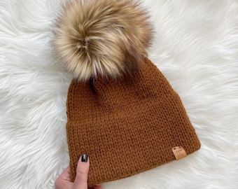 May include: A brown knitted beanie with a large faux fur pom pom on top. The beanie has a small brown leather tag with the text "Cozy & Chic" on it.