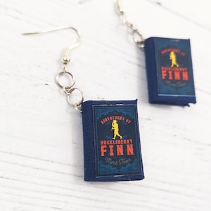 May include: A pair of earrings featuring miniature book charms with the title "Adventures of Huckleberry Finn" by Mark Twain. The charms are navy blue with red and yellow accents.