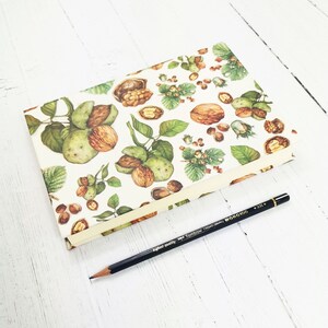 以下が含まれることがあります： 緑の葉と茶色のクルミのパターンが描かれた白いノート。ノートは閉じられており、白い木製の表面の上に置かれています。ノートの隣には黒い鉛筆が置かれています。