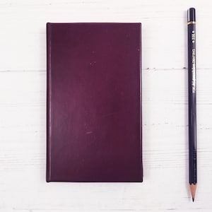 May include: A dark purple notebook with a black pencil on a white wooden surface. The pencil is labeled "FOR MECHANICAL DRAFTING 3H".