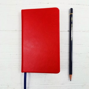 May include: A red leather-bound notebook with a blue ribbon bookmark. A black pencil is placed next to the notebook on a white wooden surface.