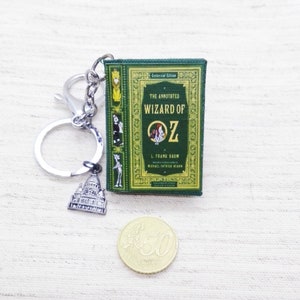 Peut inclure: Un porte-clés en forme de livre vert avec une chaîne argentée et un charme argenté représentant un bâtiment. La couverture du livre porte le texte "The Annotated Wizard of Oz" et "L. Frank Baum".