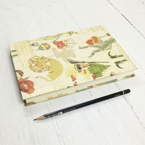 May include: A closed, cream-colored journal with a colorful, whimsical design featuring characters from the Wizard of Oz. The cover features a lion, a scarecrow, the Tin Man, and Dorothy's ruby slippers. The journal is lying on a white wooden surface with a black pencil beside it.