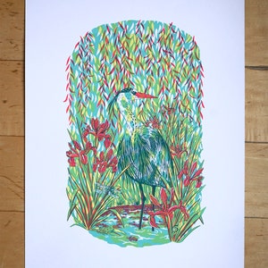 May include: A blue heron stands in a pond surrounded by green and red irises and a dragonfly. The background is a blue and green pattern of leaves.