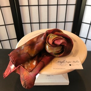 May include: A hand-dyed silk scarf in shades of brown, orange, and green, folded into a rose shape. The scarf is displayed on a white pedestal with a business card that reads "Maple Sugar Studio".