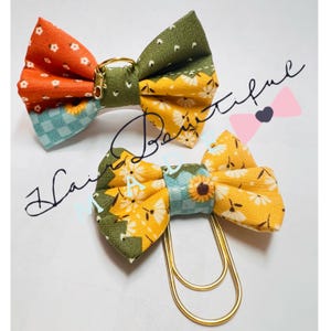 Puede incluir: Dos lazos para el pelo de tela con estampados florales y geométricos. Uno tiene una combinación de colores naranja y verde con un anillo dorado. El otro tiene una combinación de colores amarillo y azul con un clip dorado. El texto "Hair Beautiful" está escrito en cursiva.