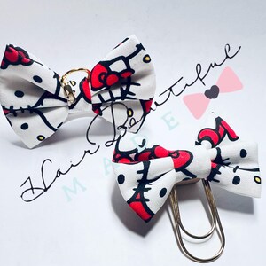 May include: Two white fabric hair bows with a Hello Kitty print. The bows have red accents and black polka dots. One bow has a gold ring attached to it, and the other bow has a gold clip.
