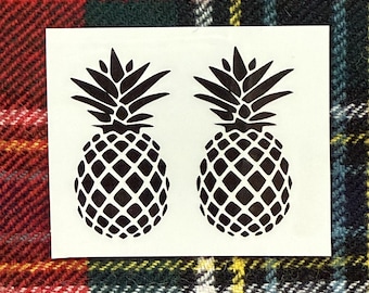 May include: Two black pineapple stickers on a white background. The pineapples have a detailed, geometric pattern and spiky tops. The stickers are placed on a plaid fabric with red, green, and yellow tones.