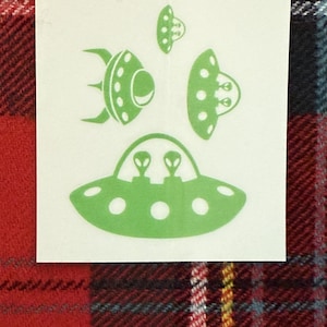 May include: A white rectangular sticker featuring green illustrations of flying saucers. The design includes several UFOs, with one showing two alien figures inside. The sticker is set against a red and black plaid background.