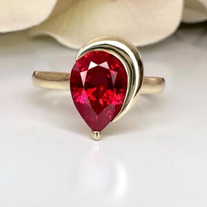 May include: A pear-shaped ruby gemstone set in a gold ring with a curved band. The ring is designed with a smooth, polished finish.