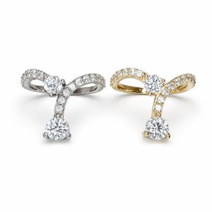 May include: Two rings, one in silver-toned metal and one in gold-toned metal, each featuring a curved band set with small clear stones and two larger round clear stones. The design includes a central prong-set stone and a dangling round stone.