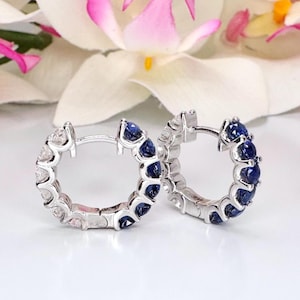 May include: A pair of silver hoop earrings with alternating white and blue gemstones. The earrings are set against a backdrop of white and pink flowers, creating a delicate and elegant presentation.