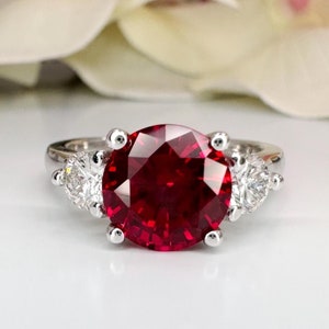 May include: A silver ring featuring a large, round, deep red gemstone as the centerpiece, flanked by two smaller, clear, round stones. The ring is set against a soft, blurred background.