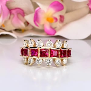 May include: A gold ring featuring a row of square-cut rubies and round diamonds. The ring has a three-tiered design with the gemstones set in a symmetrical pattern. The background includes white and pink flowers.